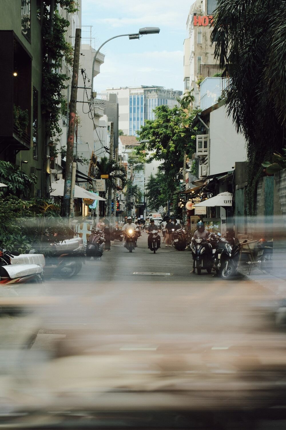 Một con phố nhỏ nhiều cây xanh ở Sài Gòn, xe máy đậu sát lề và vài chiếc chạy về phía người chụp; phần tiền cảnh nhòe vì chuyển động