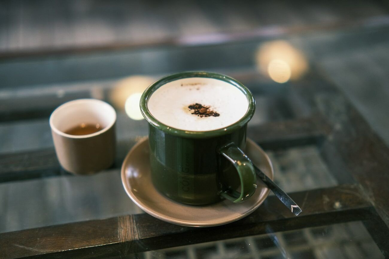 Một ly cappuccino trong cốc men xanh lá đậm đặt trên bàn kính ở một quán cà phê tại Đà Nẵng, bên cạnh là chén trà nhỏ